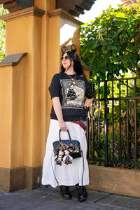 Woman wearing a graphic t-shirt and white skirt with a handbag, standing in front of a yellow wall and black metal gate.
